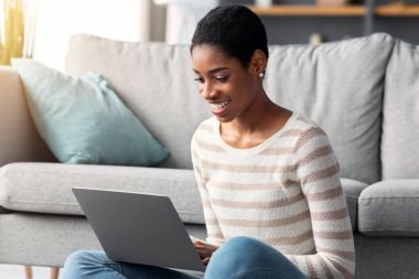Uzaktan kumanda. Gülümseyen, evde dizüstü bilgisayarla çalışan, mutlu Afrika kökenli Afrikalı Amerikalı kadın, bilgisayarda yazan, oturma odasında oturan, çevrimiçi işin tadını çıkaran, ücretsiz alan...
