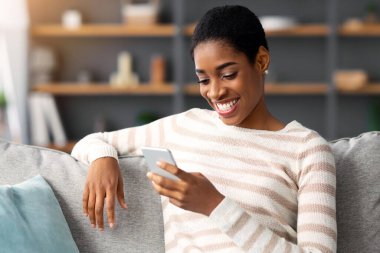 Gülümseyen siyah kadın evde dinlenirken Smartphone 'da yeni uygulamalara bakıyor, cep telefonuyla rahatlayan genç Afrikalı Amerikalı bayan, rahat oturma odasında koltukta oturuyor,