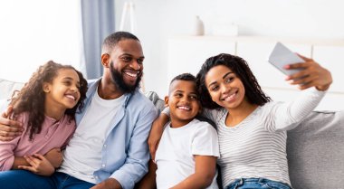 Bağlanma ve Yaşam Tarzı. Afro-Amerikan ailesinin evde selfie çekerken portresi. Mutlu ebeveynler çocuklarıyla poz veriyor ve kameraya gülümsüyor oturma odasında kanepede oturuyor.