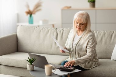 Bütçeleme, Covid-19 salgını sırasında internet üzerinden faturaları ödeme. Oturma odasındaki kanepede oturan ve aile vergilerini hesaplayan belgeler ve dizüstü bilgisayarla çalışan odaklanmış olgun bir kadının portresi.