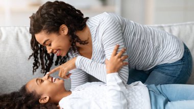 Siyah, genç bir anne ve kızı sevmek birlikte eğlenmek, ev içi, yan manzara, yakın çekim. Mutlu Afro-Amerikan annesi, kanepede birbirine yapışıp burnuna dokunarak gülen kızı gıdıklıyor.