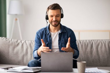 Mutlu, sakallı ve sakallı bir adam evden video görüşmesi yapıyor. Oturma odasında otururken kulaklık ve laptop kullanıyor. İş arkadaşlarıyla konuşuyor, gülümsüyor ve el kol hareketi yapıyor.