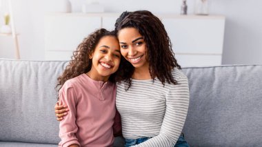 Bağ ve Birliktelik Konsepti. Afrika kökenli Amerikalı anne ve kızın portresi sarılıp gülümsüyor, birlikte vakit geçiriyorlar. Mutlu bir kadın bir kıza sarılıyor, oturma odasında kanepede oturuyor.