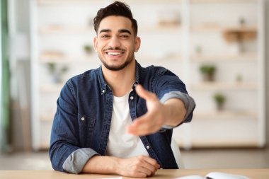 Pozitif Arap adam çalışma masasında oturuyor, kameraya gülümsüyor, ofis içi, fotokopi odası. Orta doğulu mutlu genç adam masada oturuyor, uzanmış elleriyle birini selamlıyor.