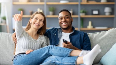 Irklar Arası Aşıklar Oturma odasında birlikte vakit geçirip televizyon izledikleri için mutlu. Neşeli, çok kültürlü çift evdeki rahat koltukta dinleniyor, televizyonu işaret eden kadın, yakın çekim...