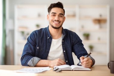 Keyifli bir Arap adam çalışma masasında oturuyor, notlar alıyor ve kameraya gülümsüyor, ofis içi, fotokopi odası. Orta Doğulu mutlu bir genç adam masada oturuyor, gazete okuyor, ev işleri yapıyor.