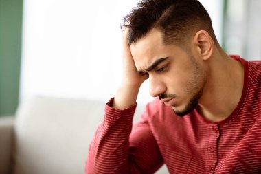 Üzgün sakallı Arap adamın yakın plan portresi. Evde koltukta oturuyor, kafasına dokunuyor ve fotokopi odası olan manzaraya bakıyor. Finansal akşamdan kalma konsepti