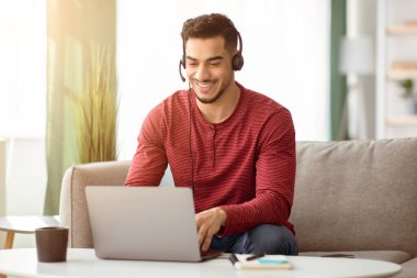 Mutlu Arap adam evde otururken internete bağlanıyor, dizüstü bilgisayar ve kulaklık kullanıyor, bilgisayar klavyesinde daktilo kullanıyor, oturma odasında kanepede oturuyor, boşluğu kopyalıyor. E-eğitim, çevrimiçi kurs kavramı