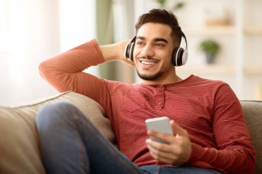 Neşeli Arap adam evdeki koltukta dinlenirken cep telefonundan müzik dinliyor, kablosuz stereo kulaklık kullanıyor, fotokopi alanına bakıyor. Neşeli orta doğulu adam akıllı telefondan müzik dinliyor.