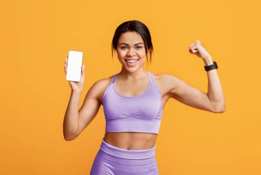 Sağlıklı yaşam tarzı ve spor uygulaması. Mutlu siyahi kadın akıllı telefonu boş ekranla gösteriyor ve sarı arka planda kol kaslarını gösteriyor. Tasarımın için bir model