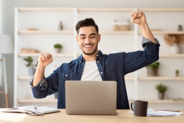Duygusal bin yıllık Arap serbest çalışanı başarılı iş anlaşmasını kutluyor, çalışma masasında dizüstü bilgisayarla oturuyor ve yumruğunu kaldırıyor, kopyalama alanı, ofis içi. Ortadoğulu adam evden çalışıyor.