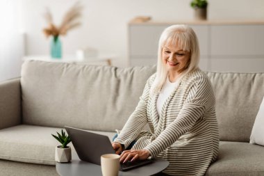 Teknoloji ve Yaşlılar. Evde dizüstü bilgisayarda çalışan, oturma odasındaki çay masasında klavyede oturan gülümseyen yaşlı kadın portresi. Olgun Kadın İnternet Tarıyor ve Öğreniyor