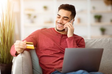 Kredi kartı tutan neşeli Arap adam, satışlarla veya banka temsilcisiyle telefon görüşmesi yapan, dizüstü bilgisayarlı koltukta oturan, ev içi, fotokopi odası olan. İnternetten ödeme yapan mutlu orta doğulu genç adam.