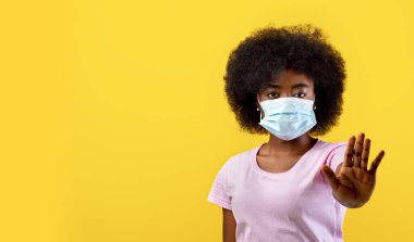Afrika kökenli Amerikalı kadın hasta, maske takıyor ve sosyal mesafeye ayak uyduruyor, sarı arka planda duruyor, stüdyo çekimi. Koruyucu maskeli siyahi kadın, el örgüsüyle çevrilmiş.
