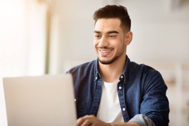 Ofiste dizüstü bilgisayarla çalışan mutlu Arap adamın yakın çekimi, defter ekranına bakıyor, neşeli bir gülümseme, kopyalama alanı. Yaratıcı orta doğulu genç adam işini seviyor. Kariyer, iş fırsatları