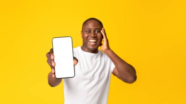 Harika haberler, zafer mesajı ve iyi bir uygulama. Beyaz tişörtlü, mutlu, Afro-Amerikalı bir adam telefonu ekranda göstermiş ve yanak yanağa bastırmış, turuncu arka planda izole edilmiş, panorama.