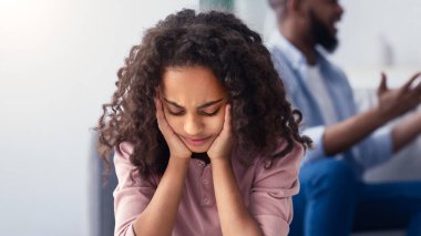 Aile içi çatışmalar. Üzgün küçük siyah kız elleriyle kulaklarını kapatırken ailesi arka planda tartışırken, üzgün çocuk kavga duymak istemiyor, stresli çocuk yalnız başına oturuyor, seçici odaklanıyor.