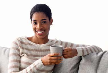 Hafta sonu eğlencesi. Oturma odasında dinlenirken sıcak içkinin tadını çıkaran ve kameraya gülümseyen Afrikalı genç bir kadın, koltukta dinlenen bir fincan kahveyle mutlu siyah güzel bir kadının portresi.