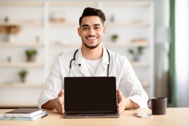 Neşeli bir ortadoğulu doktor hastanede ya da evde çalışırken bilgisayarını boş ekranla gösteriyor, kopyala, taklit et. Uzaktan sağlık hizmeti, uzak tıp konsepti