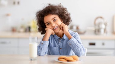 Sağlıklı Kahvaltı Konsepti. Modern mutfaktaki yemek masasında oturan tatlı, gülümseyen küçük Afrikalı Amerikalı kızın portresi. Sabahları taze süt ve kurabiyelerle.