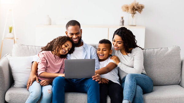 Family Weekend. Portrair of cheerful African American parents and their little children using laptop at home together, watching movie or browsing internet, sitting on the couch in living room
