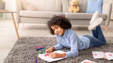 Anneler Günü. Gülümseyen kıvırcık siyah kız, annesi için kağıda kağıt çiziyor, evdeki halının üzerinde yatıyor. Kızım bayramı kutlamak için hazırlanıyor, modern iç mekan.