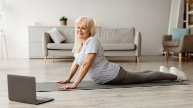 Belden aşağısı ağrıyor. Gülümseyen Olgun Kadın Sfenks Kobra Pozu Veriyor ya da Yukarı Yüzlü Köpek Asana Kaslarını esnetiyor Pilates yapıyor, Oturma odasında Yoga Mat Yapıyor, Laptop kullanıyor