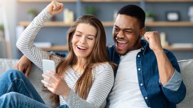 Zeki telefon ekranına bakan ve evde birlikte dinlenirken sevinçle bağıran heyecanlı ırklar arası çift, Çevrimiçi Kazananı Kutlayan Duygusal Çokkültürlü Aşıklar, Heyecanla Yumrukları Yükselten