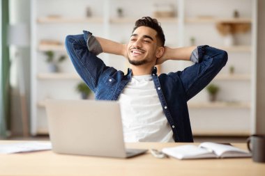 Keyifli Arap adam evden dizüstü bilgisayarla çalışırken kısa bir mola veriyor, çalışma masasında oturup arkasına yaslanıyor, gülümsüyor ve kopyalama alanına bakıyor. Çevrimiçi iş fırsatları kavramı