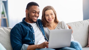 Hafta sonu eğlencesi. Oturma odasında dizüstü bilgisayarı olan ırklar arası bir çift mutlu genç, çok kültürlü aşıklar birlikte film izliyor ya da online alışveriş için bilgisayar kullanıyor.