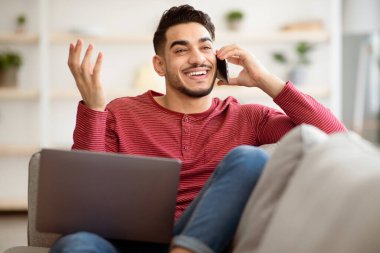 Neşeli orta doğulu adam evde takılıyor, arkadaşlarıyla telefonda konuşuyor, kanepede dizüstü bilgisayarla uzanıyor, fotokopi çekiyor. Gülümseyen Arap adam telefonla konuşuyor ve cep telefonu kullanıyor.