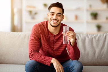 Keyifli bir Arap adam kanepeye oturmuş, su içiyor, evin içinde. Orta Doğulu genç adam elinde bardak ve gülümsemeyle evde vakit geçiriyor. Fotokopi alanı, hidrasyon konsepti.