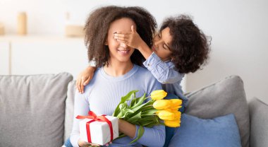 Anneye sürpriz yapıyorum. Gülümseyen siyah bir kızın portresi genç bir kadının gözlerini kapatıyor, anneler günü ya da doğum gününde karşılıyor, hediye paketini ve sarı lale buketini sunuyor.