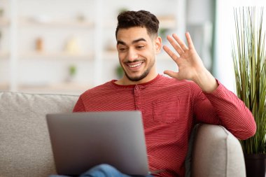 Orta doğulu mutlu bir adam dizüstü bilgisayarla kanepede oturuyor, arkadaşlarıyla online parti veriyor. Arap adam kız arkadaşıyla görüntülü görüşme yapıyor, neşeli hareketler yapıyor ve konuşuyor, boşluğu kopyalıyor.