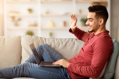 Genç, neşeli Arap adam Coronavirus salgını sırasında evde kalıp dizüstü bilgisayar ve yan görüş kullanarak video görüşmesi yapıyor. Modern teknolojiler ve iletişim konsepti