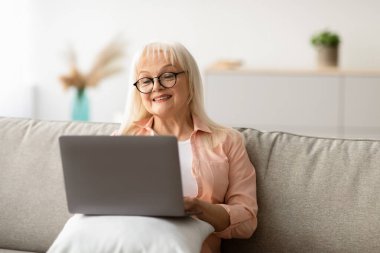 Teknoloji, Yaşlılık ve İnsanlar Konsepti. Dizüstü bilgisayar kullanarak gülümseyen, dizüstü bilgisayar başında koltukta oturan, ekrana bakan ve film izleyen son sınıf öğrencisi kadının portresi.