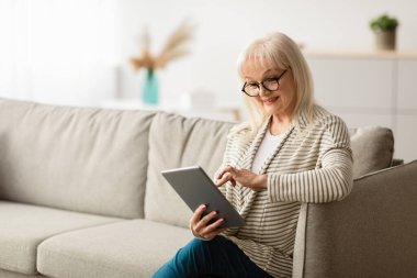 Eğlence Konsepti. Gözlüklü, elinde dijital tablet, gazete ya da e-kitap okuyan, koltukta oturan gülümseyen olgun kadın. Neşeli kadın web sayfaları, çevrimiçi video izleme, ücretsiz kopya alanı