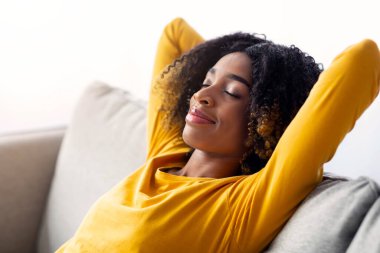 Evdeki rahat koltukta dinlenen sakin siyah kadın portresi, arkası kapalı gözlerle ve ellerle kanepeye yaslanan mutlu Afrika kökenli Amerikalı kadın, boş alanla yakın çekim.