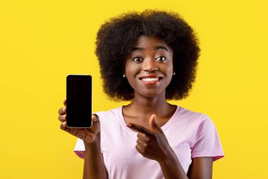 Moda cep telefonu. Heyecanlı Afrikalı Amerikalı kadın elinde siyah boş ekranla akıllı telefonu tutuyor, sarı arka planda parmağıyla işaret ediyor. Boş boşluklu Gadget, model