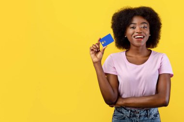 Kolay ödeme konsepti. Elinde bankamatik kartı tutan, gülümseyen ve kameraya gösteren mutlu genç siyahi kadın sarı stüdyo arka planında izole edilmiş poz veriyor.