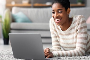 Neşeli Afrikalı Amerikalı kadın evde dizüstü bilgisayarla dinleniyor, pozitif siyah kadın oturma odasında halıda yatarken bilgisayar kullanıyor, gülümseyen kadın online alışveriş yapıyor ya da internete bakıyor.