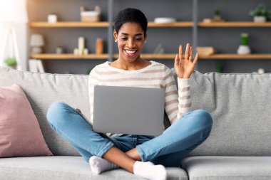 Neşeli, Afro-Amerikalı bir kadın, evinde dizüstü bilgisayarla video görüşmesi yapıyor. Dost canlısı siyahi bir kadın, oturma odasında kanepede oturmuş internet iletişiminin keyfini çıkarırken, web kamerasına el sallıyor.