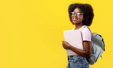 Tecritten sonra öğrenci ya da okula dönmek. Sırt çantalı ve not defterli, sarı arka planda izole edilmiş gözlüklü Afro-Amerikalı kadın.