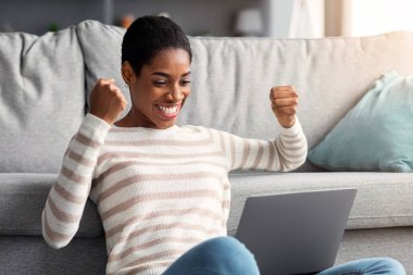 Çevrim içi Kâr. Neşeli, mutlu Afrikalı Amerikalı kadın oturma odasında otururken dizüstü bilgisayarla başarıyı kutluyor, Şanslı siyah kadın heyecanla yumruklarını kaldırıyor, boşluğu kopyalıyor.