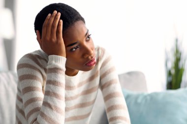 Depresyon Konsepti. Evdeki koltukta oturan üzgün Afrikalı Amerikalı kadının portresi, depresif siyah milenyum kadınının üzgün ve yalnız hissetmesi, dokunaklı kafa ve uzaklara bakması,