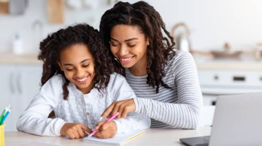 Güzel siyah anne kızına ev ödevinde yardım ediyor, mutfağın içinde. Mutlu Afrikalı Amerikalı kız dizüstü bilgisayarın önünde yazıyor ya da resim çiziyor, gülümsüyor, çocuğuna sarılıyor, boşluğu kopyalıyor.