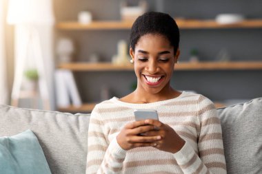 Neşeli siyahi kadın oturma odasında akıllı telefonla rahatlıyor, arkadaşlarıyla mesajlaşıyor ya da evdeki koltukta dinlenirken cep telefonuyla internete takılıyor, Afrikalı kadın mobil iletişimin keyfini çıkarıyor.