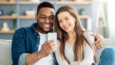 Evdeki kanepede dinlenirken akıllı telefon kullanan mutlu ırklar arası çiftler, fotoğraf izleyen neşeli çok kültürlü aşıklar ya da oturma odasında birlikte dinlenirken cep telefonlarıyla internette gezinen insanlar.