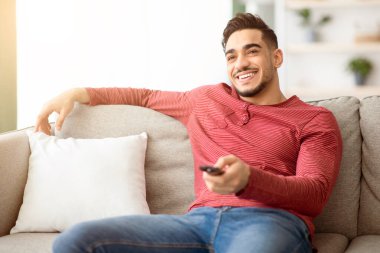 Evde TV 'de kanal değiştiren, elinde uzaktan kumandayla gülümseyen, oturma odasında kanepede oturan, televizyon izleyen, hafta sonunun keyfini çıkaran orta doğulu rahat bir adam.