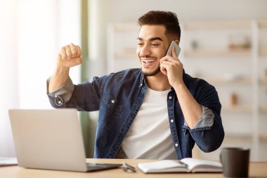 Duygusal genç Arap iş adamı ya da serbest çalışan evde dizüstü bilgisayarla çalışırken müşterisiyle ya da ortağıyla telefon görüşmesi yapan, gülümseyen ve yumruk kaldıran, defter ekranına bakan, boşluğu kopyalayan